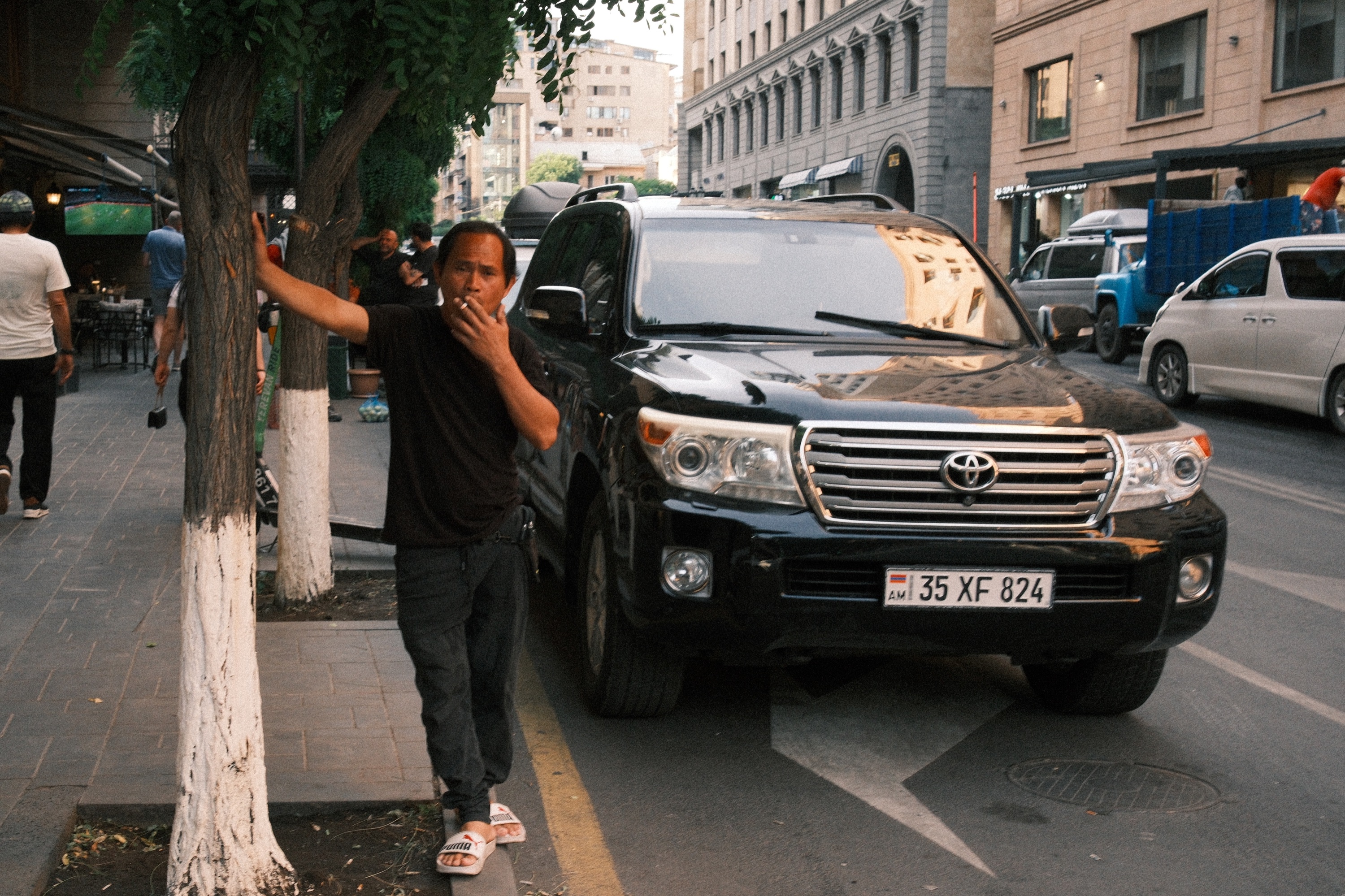 Мужчина курит, прислонившись к дереву. Рядом припаркован Land Cruiser.